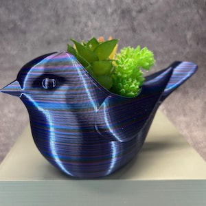May include: A blue bird-shaped planter with a textured surface, holding green and yellow artificial plants. The planter has a metallic sheen and is sitting on a light-colored surface.