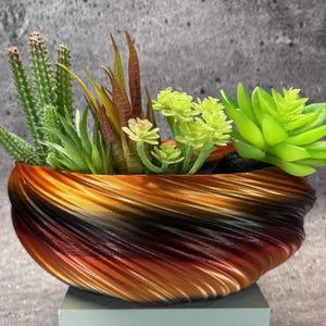 May include: A decorative planter with a swirling, textured design in shades of brown, orange, and black. The planter is filled with a variety of artificial succulents and cacti in shades of green and brown, creating a vibrant display.