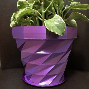 Puede incluir: Una maceta geométrica morada con una planta verde frondosa en su interior. La maceta tiene un patrón texturizado en forma de diamante.