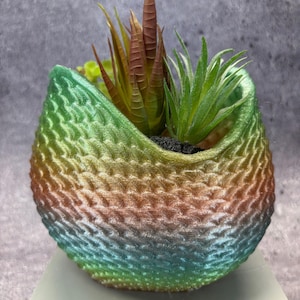 May include: A small, decorative planter with a textured, multi-colored exterior. The colors transition from green to blue, orange, and yellow. The planter holds several faux succulents with green and brown leaves. The planter sits on a light gray surface.