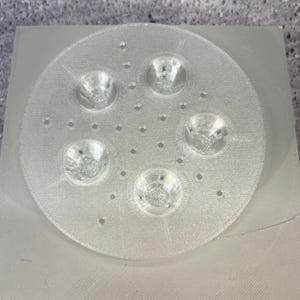 May include: A clear, circular plastic object with five recessed, bowl-shaped features and numerous small holes. The object is set on a rectangular base, both appearing to be made of the same translucent material. The design suggests a functional or decorative item.