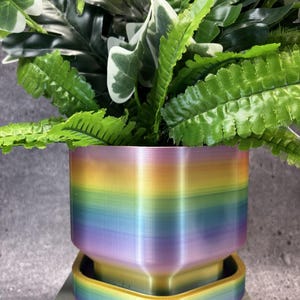 May include: A cylindrical plant pot with a smooth, gradient rainbow finish featuring horizontal bands of red, orange, yellow, green, blue, and purple. The pot sits on a matching rainbow-colored, square-shaped saucer with rounded corners.