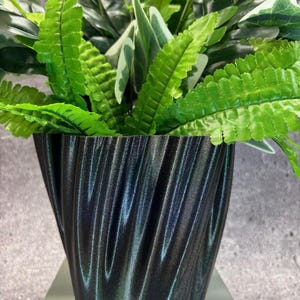 Puede incluir: Una maceta decorativa negra y texturizada con un diseño ondulado, llena de vibrantes plantas artificiales verdes. Las hojas tienen una mezcla de variegación verde y blanca, creando un aspecto natural. La maceta está sobre una superficie de color claro.
