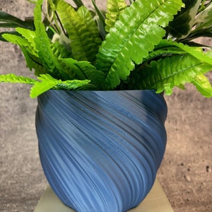 May include: A blue, textured, swirl-patterned ceramic planter with a green leafy plant inside. The planter is sitting on a light green pedestal.