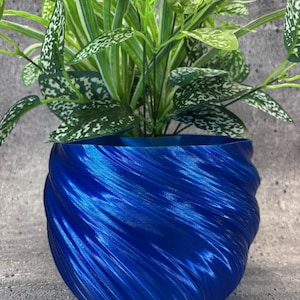 Puede incluir: Una maceta azul vibrante, con textura en espiral, que contiene una exuberante planta verde con hojas moteadas de blanco. El diseño de la maceta presenta un patrón fluido en forma de ola, creando un aspecto moderno y elegante.