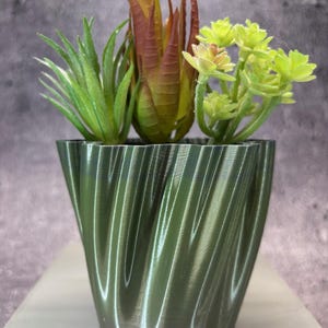 Op de afbeelding: Een decoratieve groene plantenbak met een wervelend ontwerp, met drie kunstmatige vetplanten. De vetplanten hebben groene, bruine en gele tinten, wat een natuurlijke uitstraling creëert. De plantenbak is ongeveer 13 cm hoog.