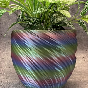 Puede incluir: Un macetero de espiral verde, azul y morado con una planta dentro. El macetero está hecho de un material liso y brillante.
