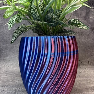 May include: A cylindrical plant pot with a spiralling, ribbed texture featuring a metallic colour-shifting finish that transitions from deep blue to vibrant magenta. A green and white variegated houseplant is potted inside the container.