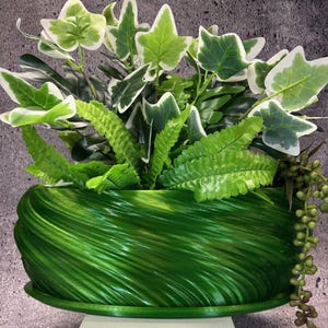 Op de afbeelding: Een groene plantenbak met een draaiend, getextureerd ontwerp. De plantenbak is gevuld met verschillende groene planten, waaronder klimop en varens.