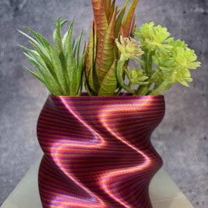 Puede incluir: Una maceta decorativa rojo-violeta, impresa en 3D, con un diseño ondulado y acanalado. La maceta contiene una variedad de suculentas artificiales en tonos verdes, amarillos y marrones. La maceta mide aproximadamente 13 cm de alto.