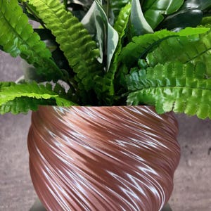 May include: A rose gold swirl planter with a green leafy plant inside. The planter is made of a smooth, shiny material and has a unique, swirling design.