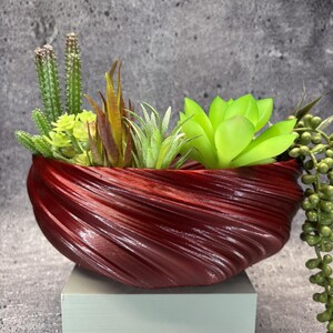 May include: A deep red, bowl-shaped planter with a textured, swirling ribbed surface. It contains an arrangement of various artificial succulents, including tall green cacti, spiky air plants, a bright lime green rosette, and trailing beads.