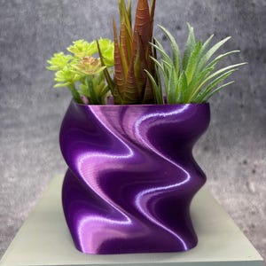May include: A glossy, purple, wave-patterned planter holding artificial succulents. The planter is on a light green surface. The faux plants feature green, brown, and light green leaves. The planter's dimensions are approximately 15 cm in height.