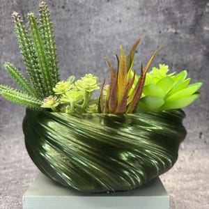 Op de afbeelding: Een donkergroene, metallic kom met een golvende, geribbelde textuur gevuld met kunstmatige vetplanten. De planten bestaan uit hoge, stekelige groene cactussen, lichtgroene bladstengels en een centrale groep puntige, roodbruine bladeren.