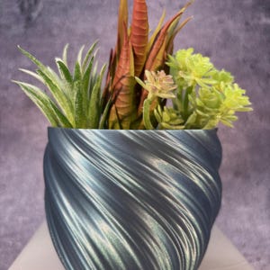 Op de afbeelding: Een teal blauwe swirl-pot met een verscheidenheid aan kunstmatige vetplanten. De vetplanten zijn groen, rood en oranje.