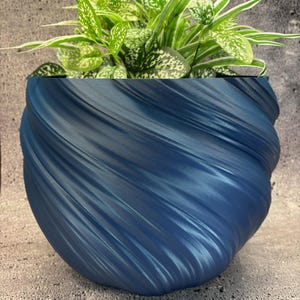 Puede incluir: Una maceta decorativa azul, texturizada, con un diseño en espiral, llena de una planta de hojas verdes y blancas. La maceta tiene forma redondeada y se asienta sobre una superficie gris. La planta tiene hojas verdes con motas blancas.