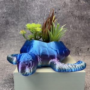 May include: A sea turtle-shaped planter with a glossy, iridescent finish that shifts between shades of blue, purple, and green. The shell serves as a container holding several small, artificial green and brown succulent plants.