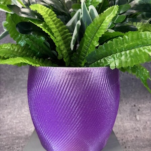 May include: A vibrant purple planter with a textured, diagonal stripe design, filled with lush green and white artificial plants. The planter sits on a light gray surface, creating a striking contrast.
