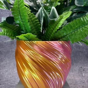 Puede incluir: Un arreglo de plantas decorativo con exuberante follaje verde en una maceta metálica con remolinos. La maceta tiene un degradado de color de rosa a dorado. La planta incluye varias formas y texturas de hojas.