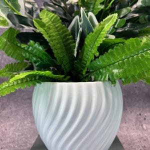 May include: A white, round plant pot with a textured, swirling design, filled with vibrant green and white artificial plants. The faux foliage includes ferns and other leafy varieties, creating a decorative accent.