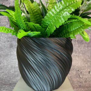 May include: A black, textured, spiral-patterned planter filled with vibrant green artificial ferns and variegated leaves. The planter sits on a light-colored, square pedestal, creating a modern, decorative display for home or office.