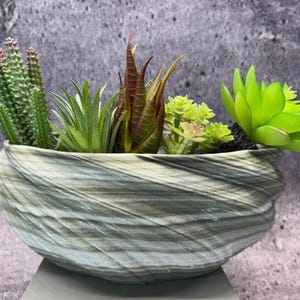 Peut inclure: Un pot décoratif en céramique avec un motif tourbillonnant texturé dans des tons de gris et de blanc. Le pot est rempli d'une variété de succulentes et de cactus artificiels verts, bruns et rouges. Un élément de décoration intérieure moderne.