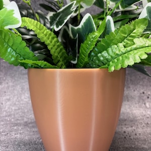 May include: A light brown ceramic planter with a smooth, textured surface. The planter is filled with green leafy plants.