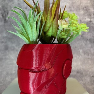 Puede incluir: Una maceta roja con forma de casco de Iron Man, impresa en 3D, llena de suculentas artificiales variadas. La maceta tiene un acabado brillante y un diseño detallado. Las plantas artificiales presentan tonos verdes, amarillos y marrones.