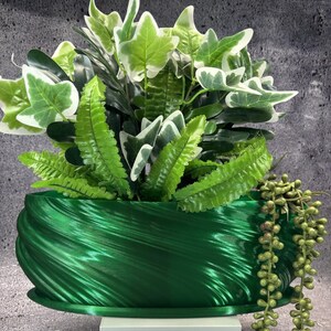 May include: A metallic emerald green oval planter with a swirling, ribbed texture. The planter is filled with an arrangement of artificial greenery, including variegated ivy leaves, bright green fern fronds, and a trailing string of pearls plant.