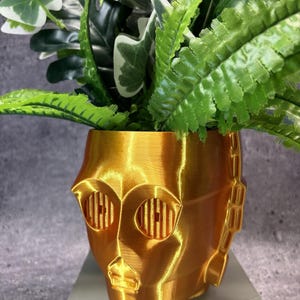 May include: A metallic gold 3D-printed planter shaped like a robotic head with circular eye cutouts and a structured jawline. The planter is filled with a variety of green artificial foliage, including fern-like leaves and broad, variegated white and green leaves.