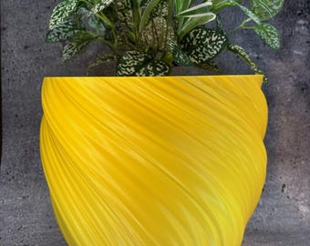 Könnte beinhalten: Ein leuchtend gelber, spiralförmig strukturierter Pflanzentopf enthält eine grüne Pflanze mit weiß gefleckten Blättern. Das Design des Topfes hat eine fließende, geschwungene Form und erzeugt eine moderne Ästhetik. Die Blätter der Pflanze haben ein gesprenkeltes Muster.