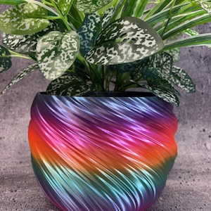 May include: A colourful planter pot with a swirling design, featuring a gradient of rainbow hues. The pot contains a green plant with white-spotted leaves. The pot's colours transition from purple to orange to teal, creating a striking contrast.
