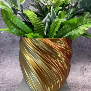 May include: A gold-colored, spiral-textured planter pot holding a variety of green and white leafy plants. The pot has a metallic sheen and sits on a light gray surface. The plants have various shades of green.