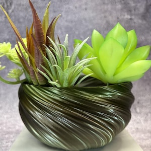 Puede incluir: Una maceta de color verde oliva oscuro con una textura acanalada en espiral que contiene una variedad de suculentas artificiales. El arreglo incluye una roseta grande de color verde lima brillante, una planta de aire espinosa central y hojas puntiagudas de color marrón rojizo.