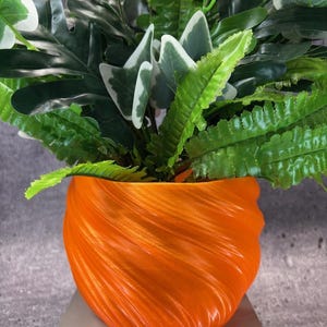 May include: A bright orange planter with a distinct spiral, ribbed texture containing a lush arrangement of artificial green foliage. The plant features a mix of broad, dark green leaves and lighter, variegated leaves with white edges.