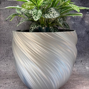 May include: A silver-colored, spiral-textured planter pot holding a green plant with white-speckled leaves. The pot has a unique, swirling design and sits on a textured surface, creating a modern aesthetic for home decor.