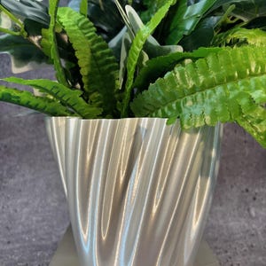 May include: A silver, wavy, textured planter with a green leafy plant inside.