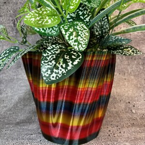 Pode incluir: Uma planta com folhas verdes e brancas em um vaso colorido, listrado, ondulado, impresso em 3D. O vaso é vermelho, amarelo, azul e preto.