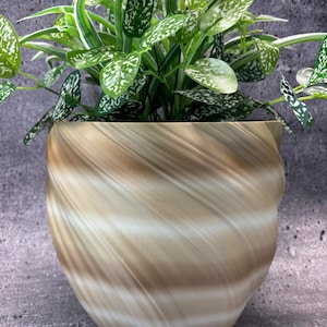 Op de afbeelding: Een decoratieve plant in een beige en bruin gedraaide plantenbak. De plant heeft groene bladeren met witte stippen en wit en groen gestreepte bladeren. De plantenbak heeft een uniek, getextureerd ontwerp.