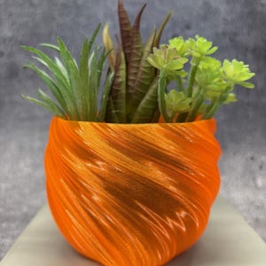 May include: An orange, spiral-textured planter filled with a variety of small, artificial succulents. The succulents include green, brown, and light green plants. The planter sits on a white surface against a gray background.