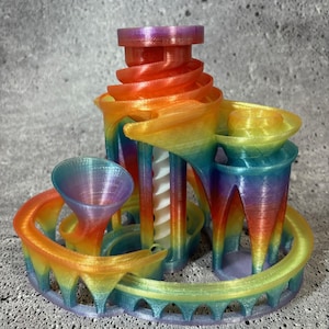 May include: A colorful, 3D-printed marble run toy in a rainbow gradient. The design features a spiral tower, curved tracks, and arches. The toy is made of plastic and has a smooth, textured finish.