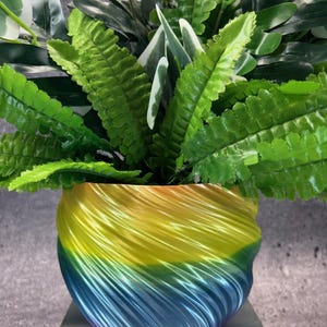 Op de afbeelding: Een levendige, regenboogkleurige bloempot met een wervelend ontwerp, met een verscheidenheid aan groene planten. De pot loopt over van geel naar blauw, wat een aantrekkelijk contrast creëert. De planten hebben verschillende tinten groen en wit.