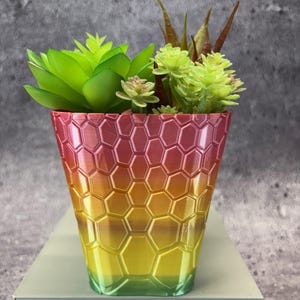 May include: A decorative planter with a honeycomb pattern, featuring a gradient of colors from red to yellow to green. The planter holds a variety of artificial succulents in shades of green and brown, creating a vibrant display. The planter is approximately 5 inches tall.