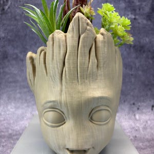 Puede incluir: Una maceta beige impresa en 3D con forma de cabeza de personaje, con una cara sonriente y ojos grandes. La maceta está llena de una variedad de plantas artificiales verdes y burdeos. La maceta está sobre una superficie gris claro.