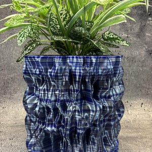 May include: A cylindrical plant pot with a deep blue and silver metallic finish, featuring a complex, rippled, and vertically pleated texture. A lush green and white variegated houseplant with pointed leaves grows out of the top of the container.