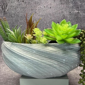 Peut inclure: Un arrangement décoratif de plantes succulentes artificielles dans un bol en céramique gris tourbillonnant. L'arrangement comprend diverses plantes vertes et brunes, avec une vigne de petites perles vertes rondes. Le bol repose sur un petit piédestal gris.