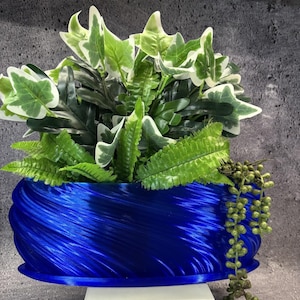May include: A vibrant royal blue planter with a distinct swirling, textured surface pattern. The planter is filled with a lush arrangement of artificial green ivy leaves with white edges, fern fronds, and a trailing string of green beads.