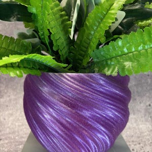 May include: A vibrant, artificial plant arrangement featuring lush green foliage and a mix of white-edged leaves. The plant is housed in a textured, swirling purple pot, creating a striking contrast. The pot sits on a light gray surface.