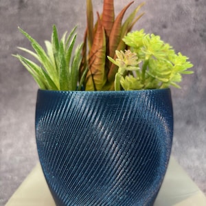 Pode incluir: Um pequeno vaso decorativo azul escuro com um design texturizado e rodopiante. O vaso contém suculentas artificiais em tons de verde, laranja e branco. O arranjo está sobre uma superfície branca.