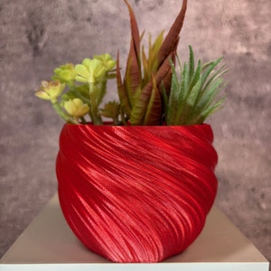 Puede incluir: Una maceta roja vibrante, texturizada con un diseño en espiral, llena de suculentas artificiales. Las plantas son una mezcla de verde, amarillo y marrón rojizo, creando un contraste colorido. La maceta está sobre una superficie blanca.